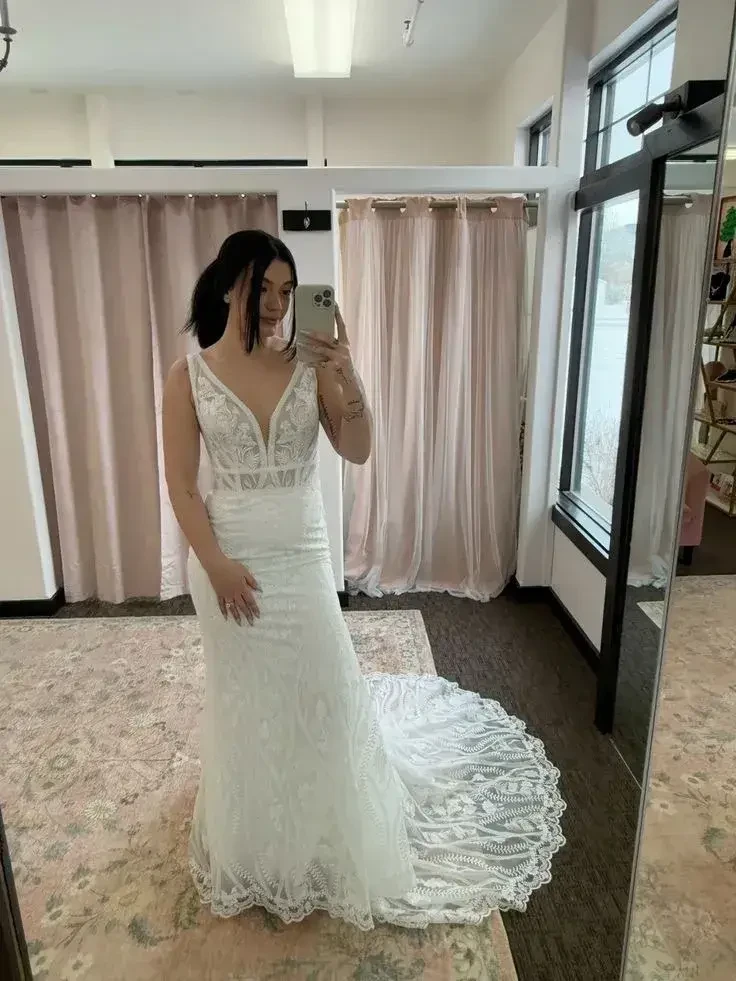 A woman takes a mirror selfie in a bridal shop, wearing a fitted, lace wedding gown with an elegant train. Soft lighting and pink curtains create a serene atmosphere.