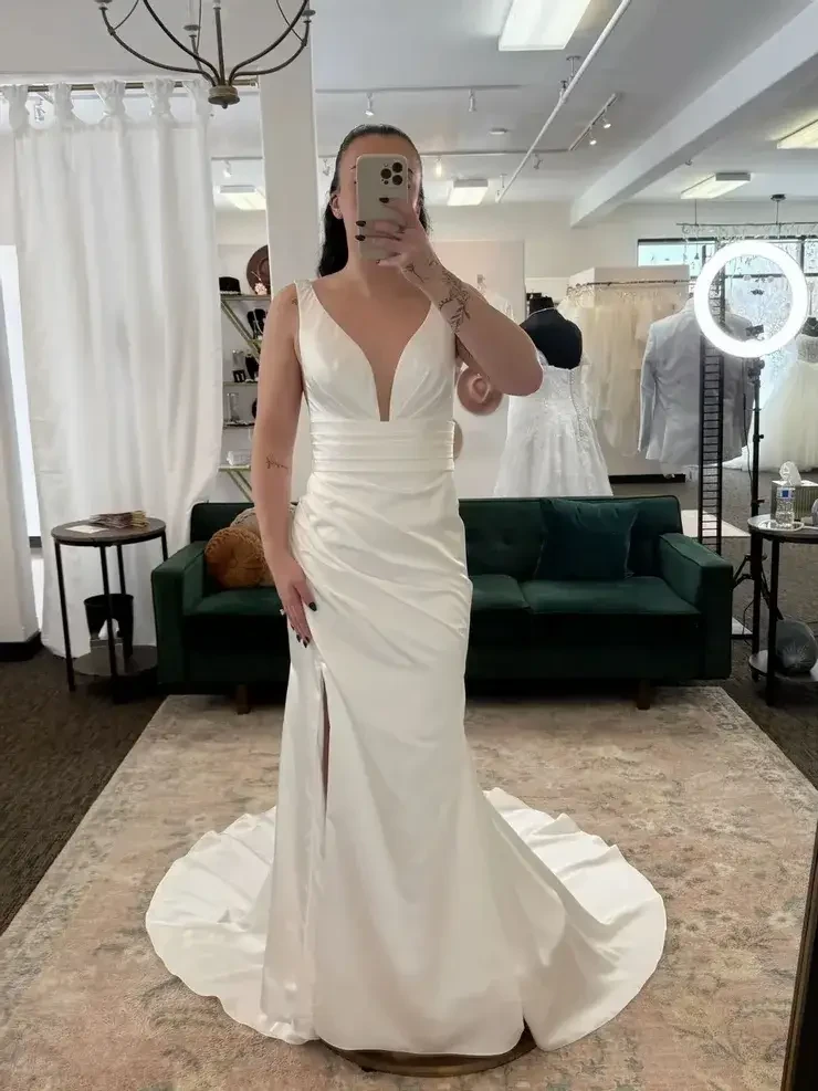 A person takes a selfie in a bridal shop wearing a white satin wedding dress with a deep neckline and side slit. The room has a cozy vibe with a green sofa, elegant rugs, and bridal gowns displayed.