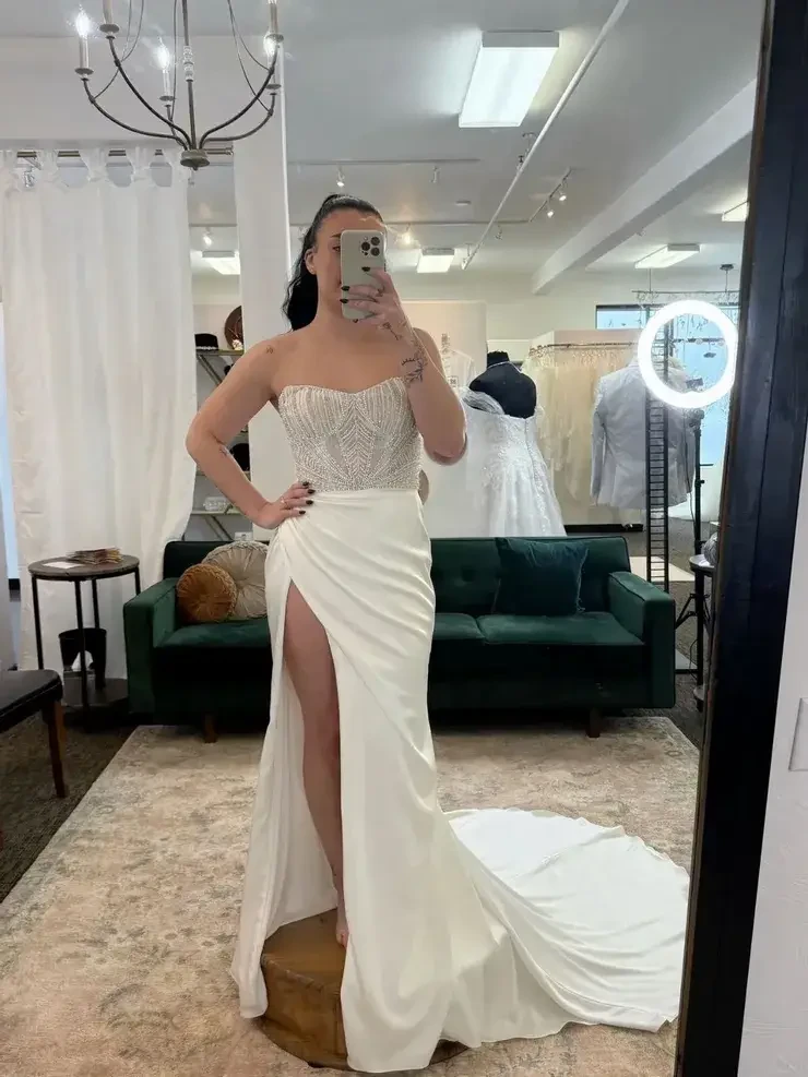 A person in a strapless wedding dress with a beaded bodice and thigh-high slit poses in a boutique. They stand on a pedestal, capturing a mirror selfie. A chandelier, green couch, and gowns are visible in the bright room, creating an elegant atmosphere.